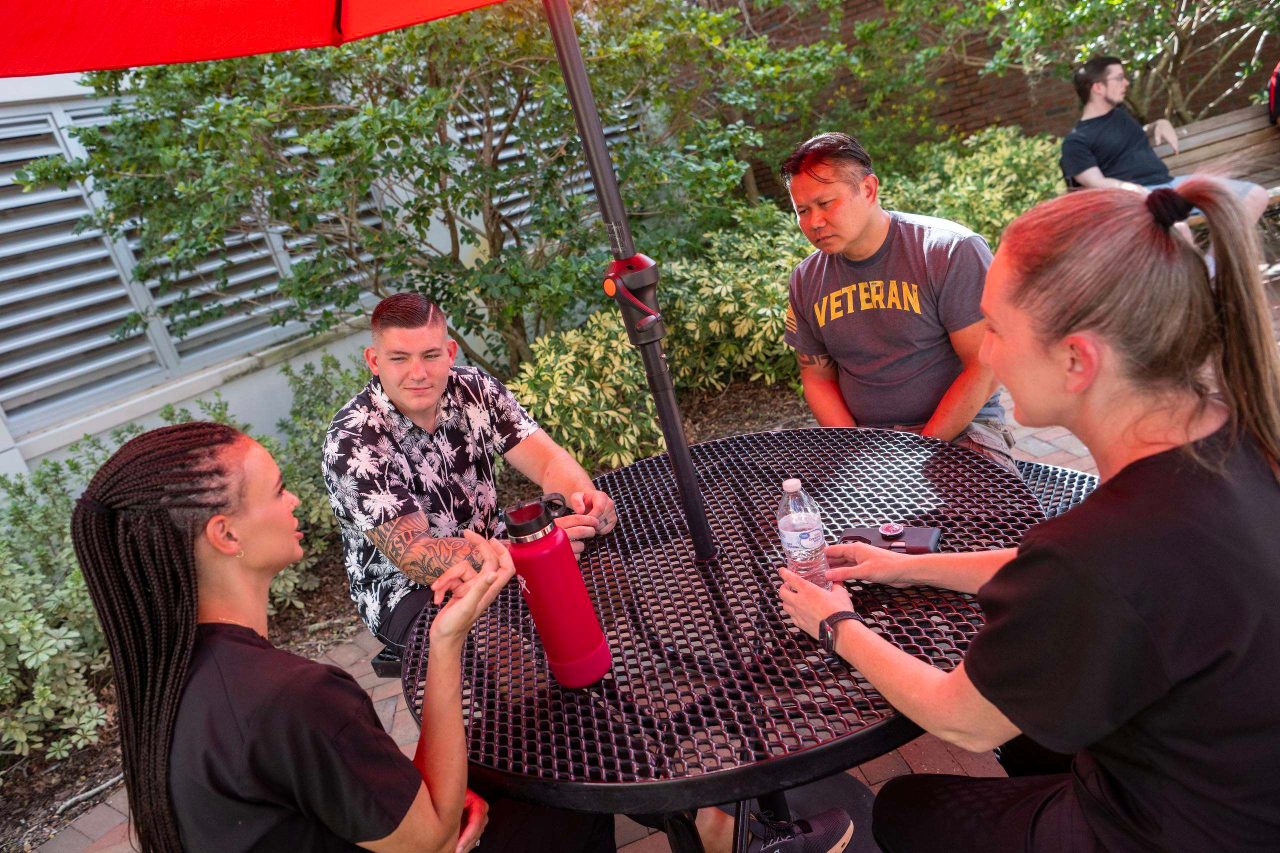 Four people sit around an outdoor patio table under a red umbrella, engaged in conversation. One student wears a shirt with the word “Veteran,” while the group appears to be participating in a casual discussion in a shaded campus courtyard surrounded by greenery.