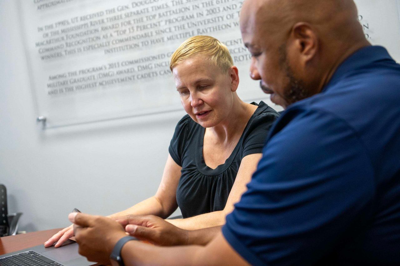 Two individuals sit together at a table reviewing information on a laptop and mobile device. The image highlights a one-on-one advising or support meeting in a professional campus setting.