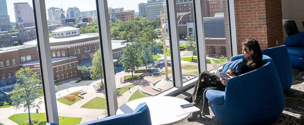 A student in the Graduate and Continuing Studies office, sitting in a chair and looking through the glass window at the College of Business.
