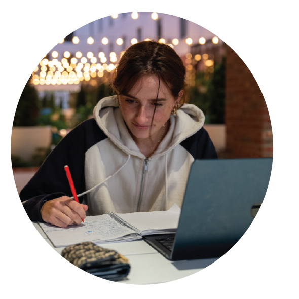 : A WOMAN IS SEATED AT A TABLE OUTSIDE. SHE IS TAKING NOTES IN A NOTEBOOK