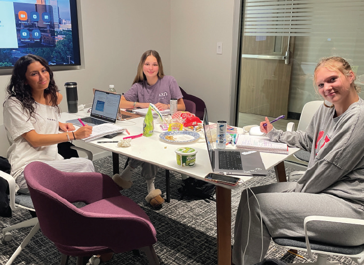 A PHOTO OF THREE WOMEN AT A TABLE. THE TABLE HOLDS LAPTOPS, FOOD AND DRINKS
