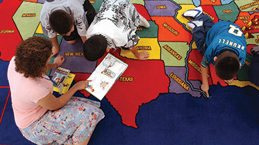 A student-teacher working with elementary students on a rug on the floor.