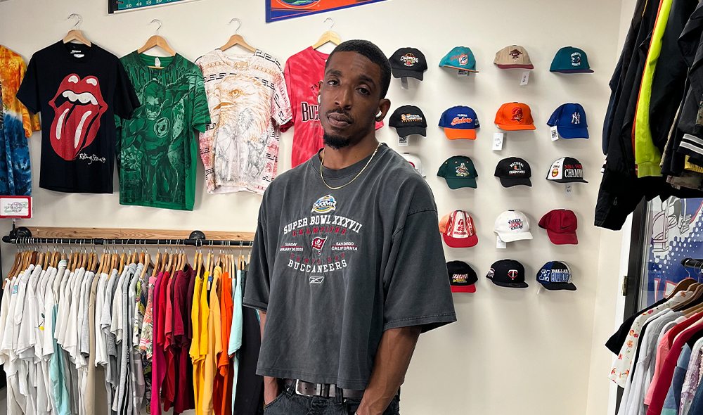 A MAN WEARING JEANS AND A T-SHIRT STANDS IN FRONT OF A WALL WITH BASEBALL CAPS DISPLAYED AND CLOTHES HANGING ON A RACK
