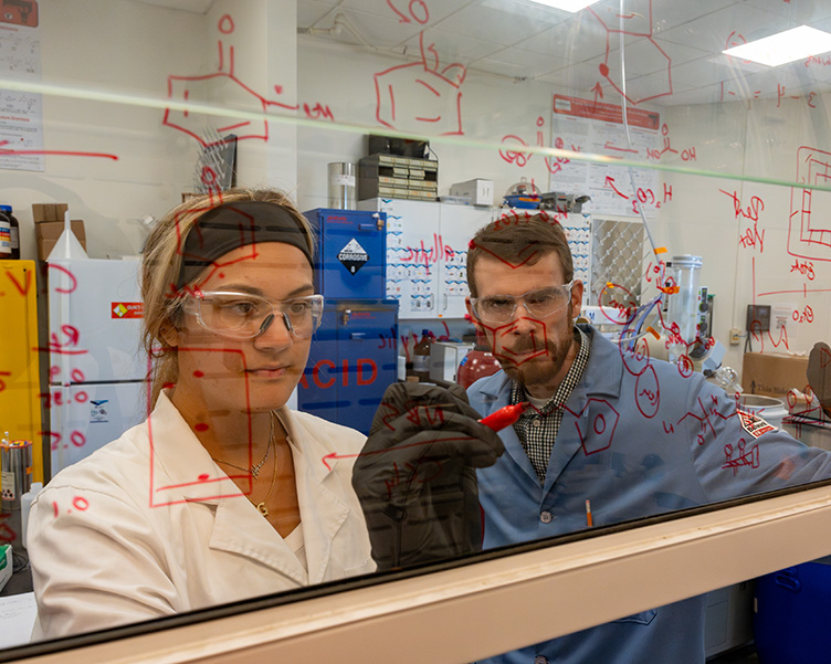 A professor and student work on chemistry calculations on a wipeboard.
