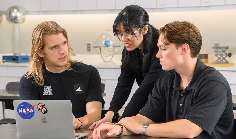 THREE PEOPLE GATHER AROUND A LAPTOP COMPUTER IN A SCIENCE LAB.