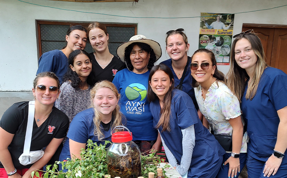 A GROUP OF 10 WOMEN POSE FOR A PICTURE. ON A TABLE ARE ARE HERBS AND LEAVES AND BOTTLES