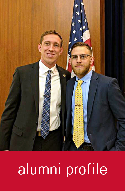 Two men stand in front of an American flag for a photo. Alumni Profile is typed in at the bottom with a red background.